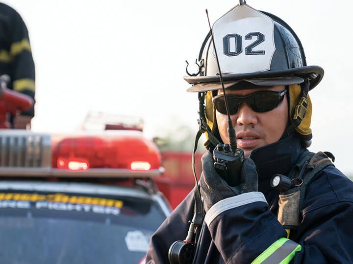 Firefighter in full protective gear and sunglasses talking on a radio in front of a fire engine