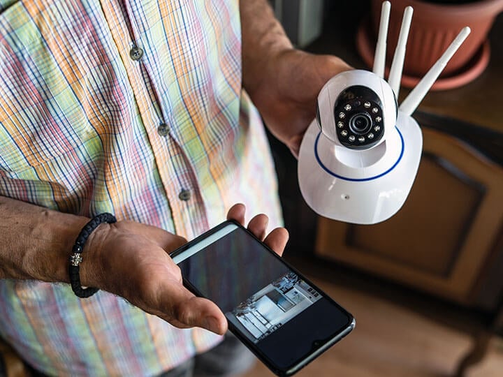 Close up of man in a multicolored button up shirt, holding a home security surveillance camera and mobile phone displaying the video feed from the camera