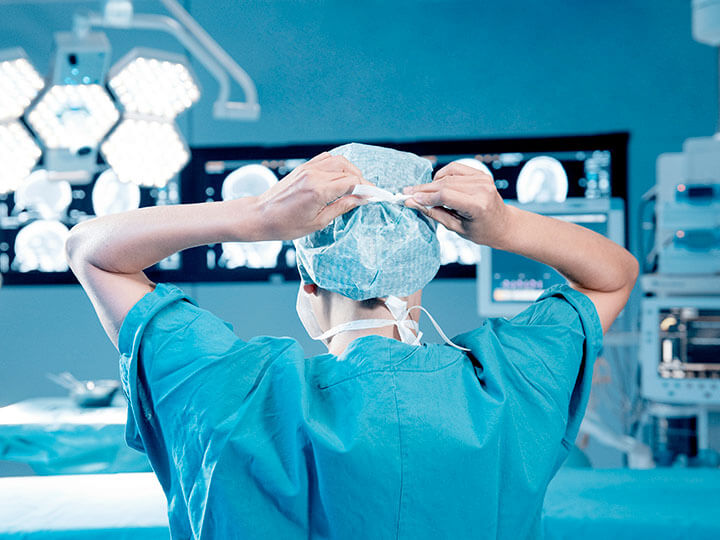 Rear view of surgeon in blue scrubs putting on a protective cap and face mask before surgery, with the operating room in the background