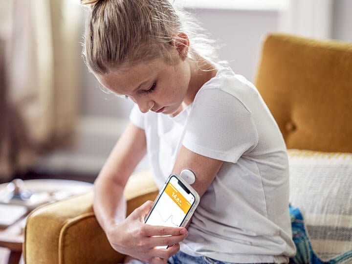 Child reading her blood sugar level with her cellphone by placing it next to her pump in her arm as she sits on her living room couch