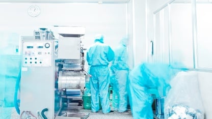 Workers in protective suits operate pharmaceutical production machinery in a bright, sterile laboratory setting.